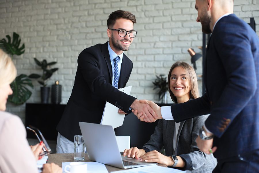 Auf dem Bild sieht man zwei Männer in Businesskleidung sich die Hand geben, während zwei Damen das Geschehen beobachten.