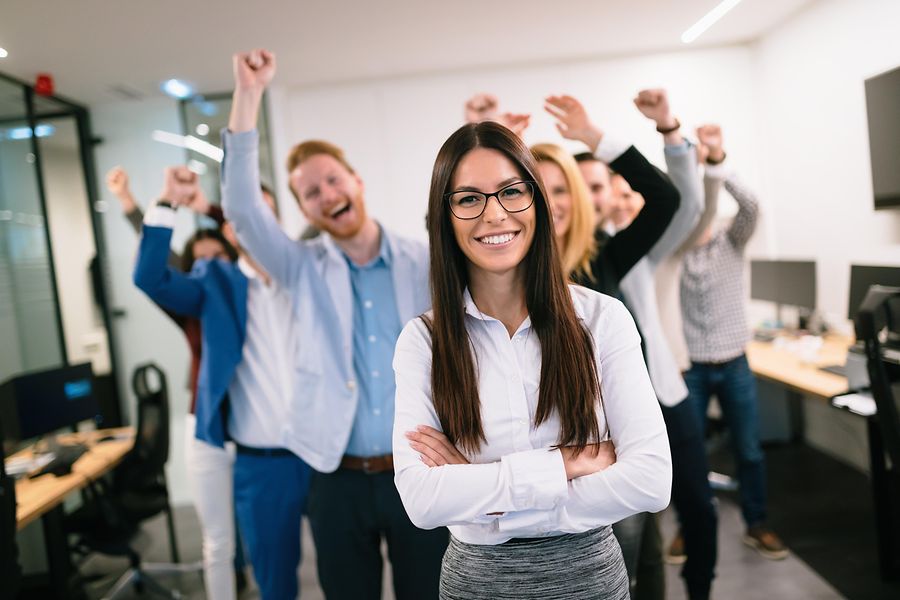 Das Foto zeigt im Vordergrund eine Geschäftsfrau. Im Hintergrund seht ihr Team, dass sich über den Erfolg freut.